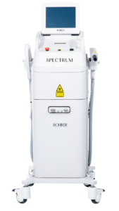 A tall, white Spectrum laser platform with a rectangular touchscreen, multiple buttons on the control panel, a yellow laser warning label, and handpiece attachments on the sides. The device sits on four caster wheels and has a smooth, glossy finish.